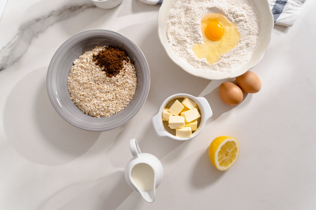 An overhead shot of baking ingredients including flour, eggs, butter, milk, and spices on a white counter.
